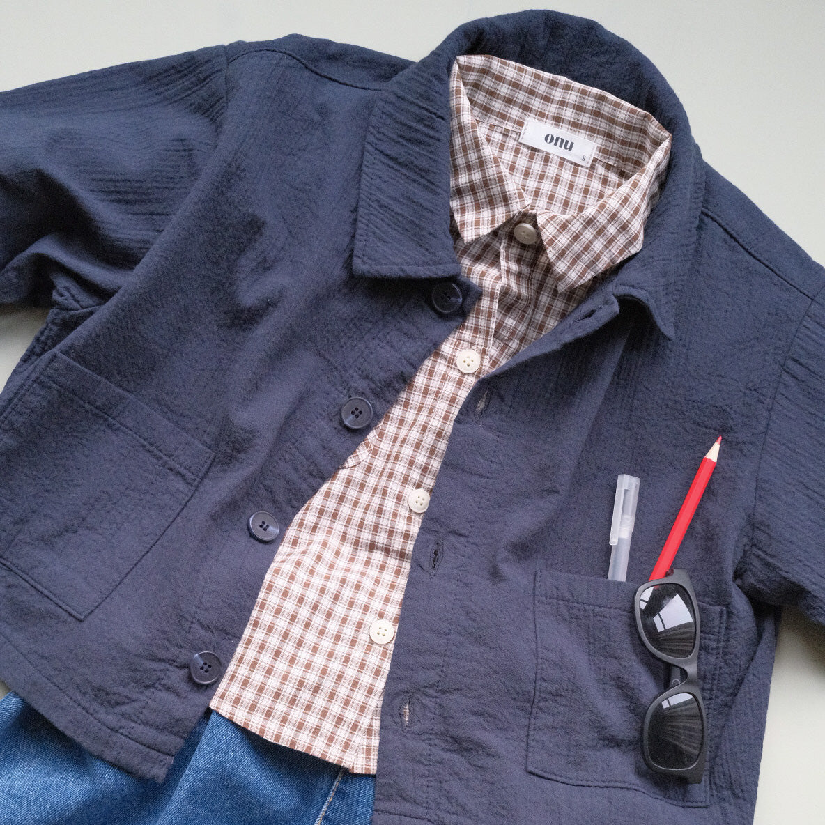 Navy blue  chore jacket with plaid brown shirt, sunglasses, and red pen on a light gray background