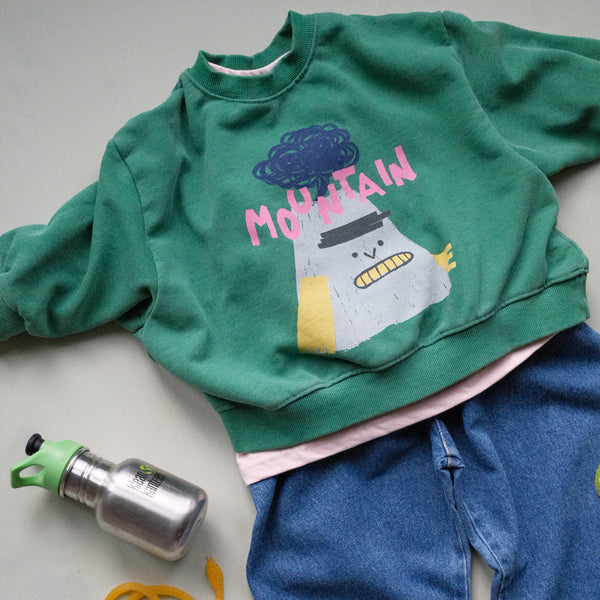 Green sweatshirt with cartoon mountain volcano design and blue barrel leg jeans on a light gray surface.