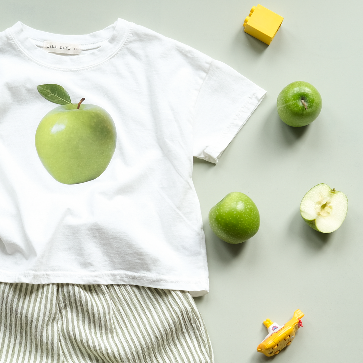 Children's outfit with white shirt and striped pants, surrounded by green apples and toys on a gray background