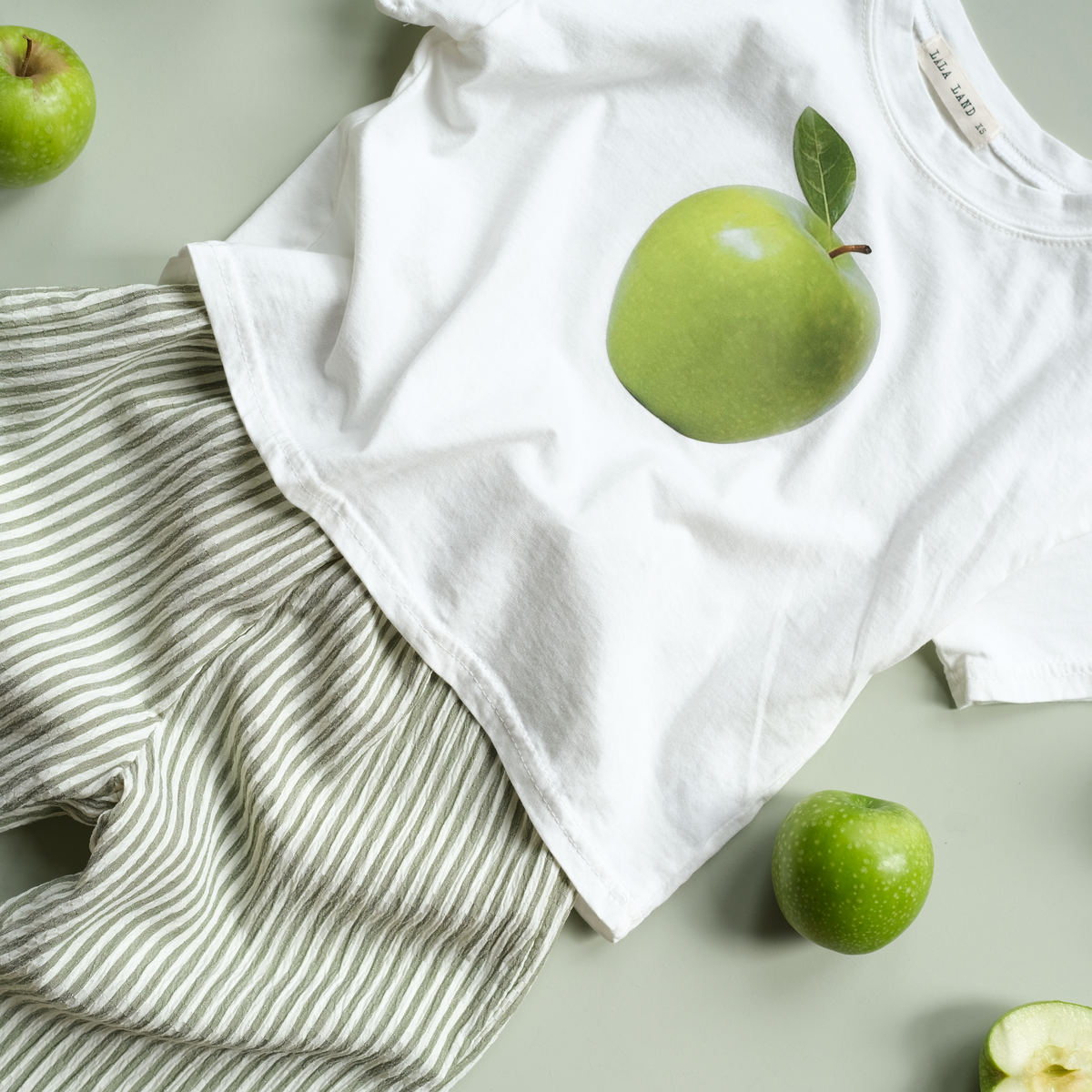 White t-shirt with green apple design and striped pants on a light gray background with green apples.